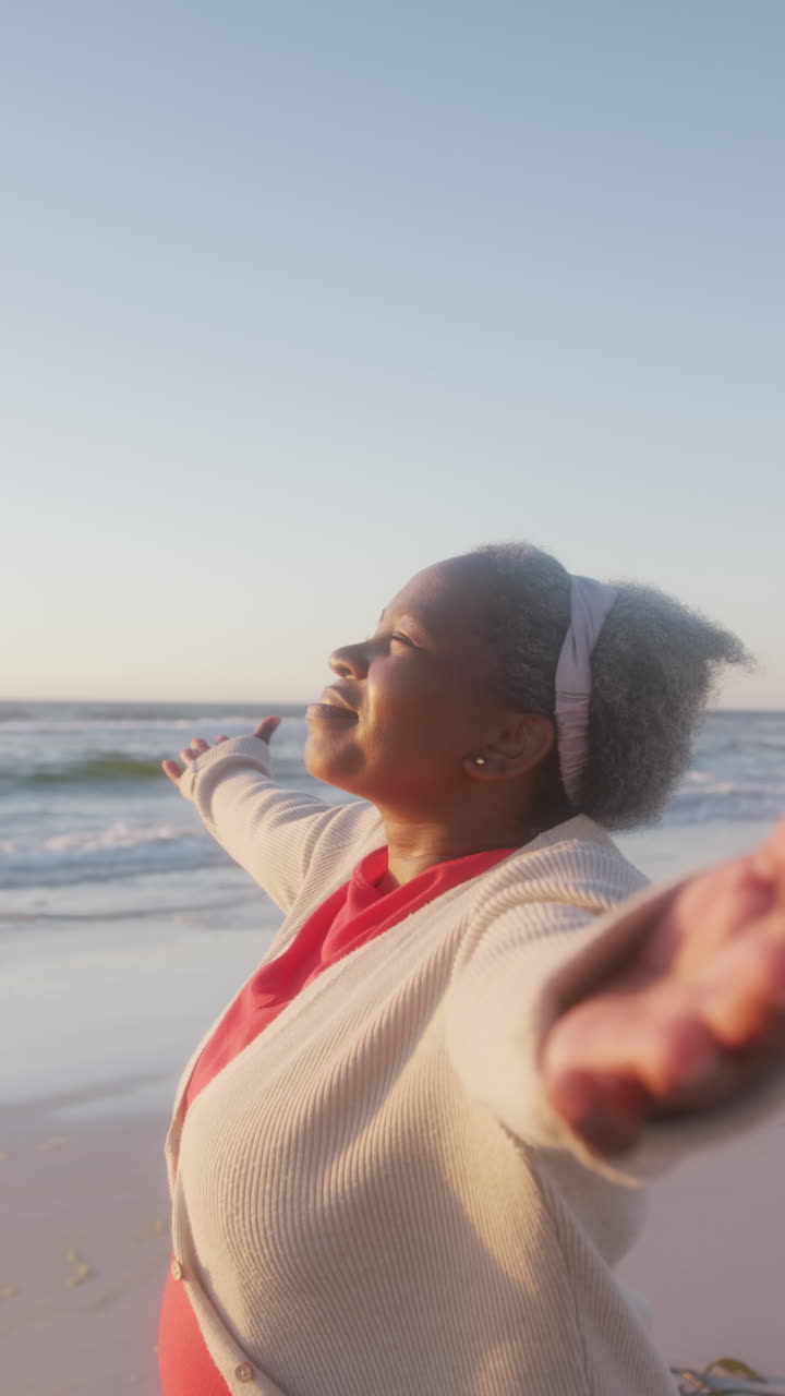 Vertical video of happy senior african american woman widening arms at beach, in slow motion