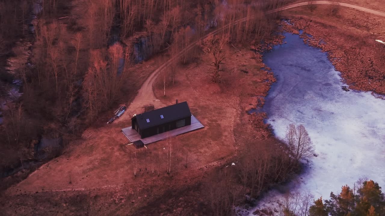 Droneoptagelser af en sort hytte omgivet af skov og en frossen sø. Filmisk naturlandskab ideelt til rejser, udendørs og baggrundsbrug.