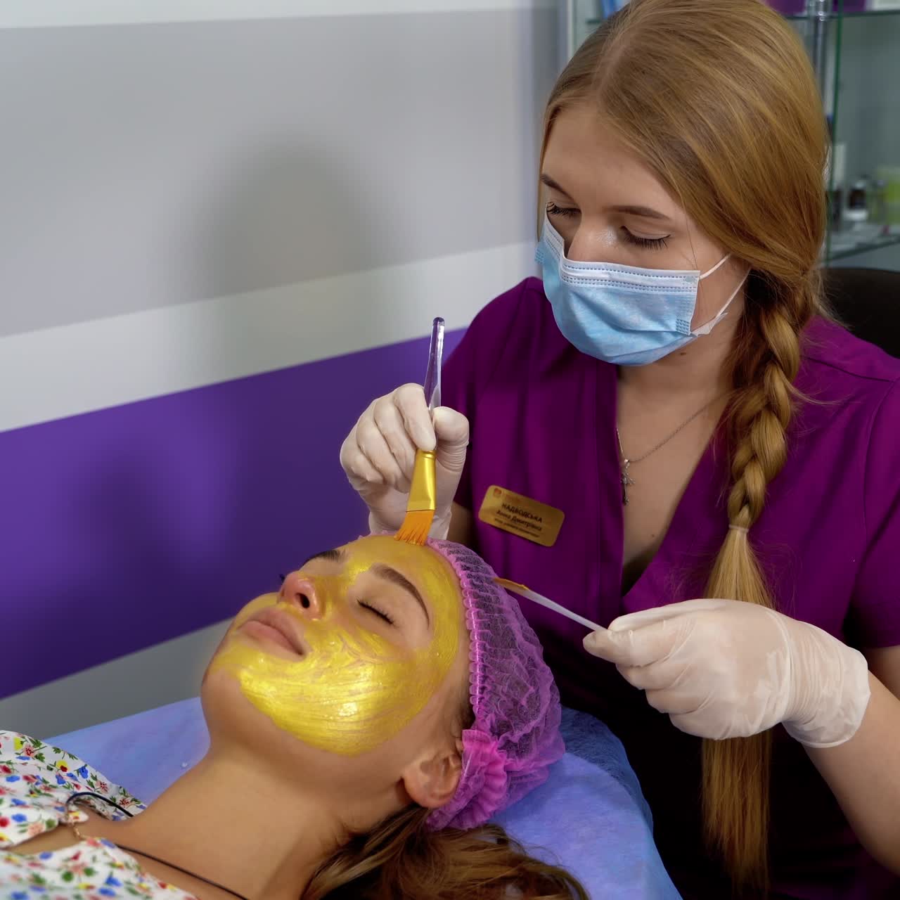 Cosmetologist apply gold mask with brush on young pretty woman`s face in beauty clinic. Spa salon. Beautician making beauty facial skincare procedure