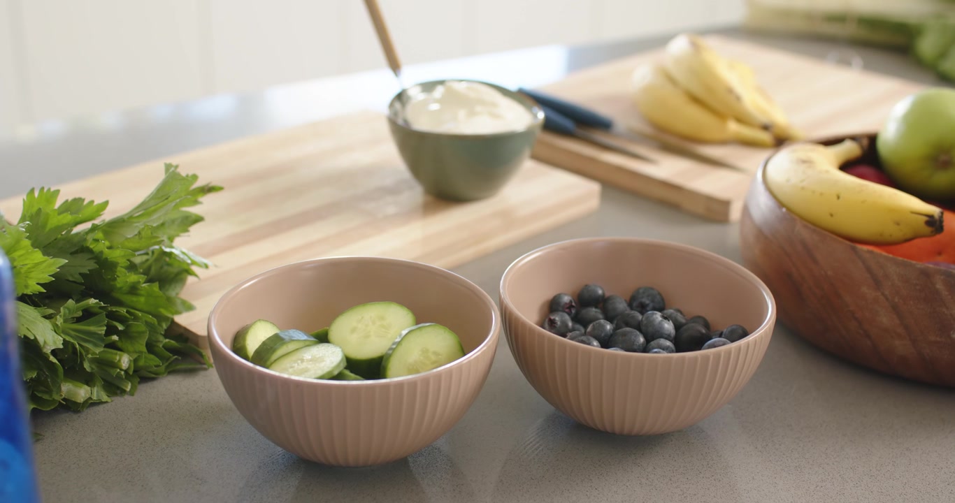 Camera is shifting focus, showing two ceramic bowls with cucumber and blueberries on counter
