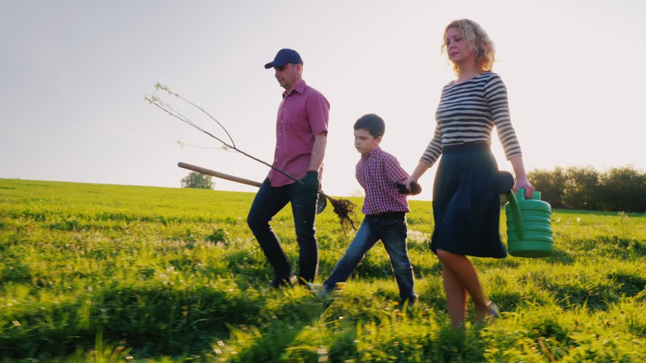 A family of farmers with a small son go together to plant a tree. Bear the apple tree seedlings, shovel and watering pad. Steadicam shot, side view