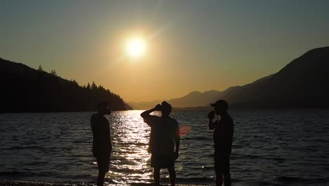 puesta de sol en un lago con montañas en el fondo con tres tipos tomando una copa en silueta