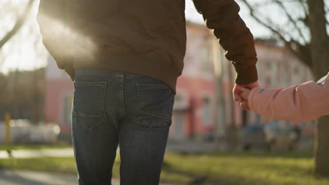 vista trasera de una niña en una gorra rosa y chaqueta, y un hombre en una chaqueta marrón, ambos con vaqueros azules, tomados de la mano mientras caminan por una calle soleada