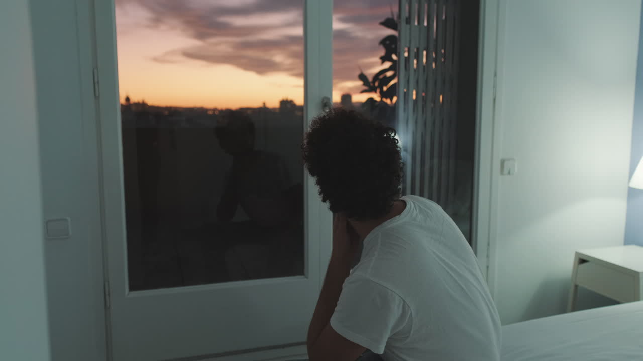 Man in bedroom looking at sunset through window