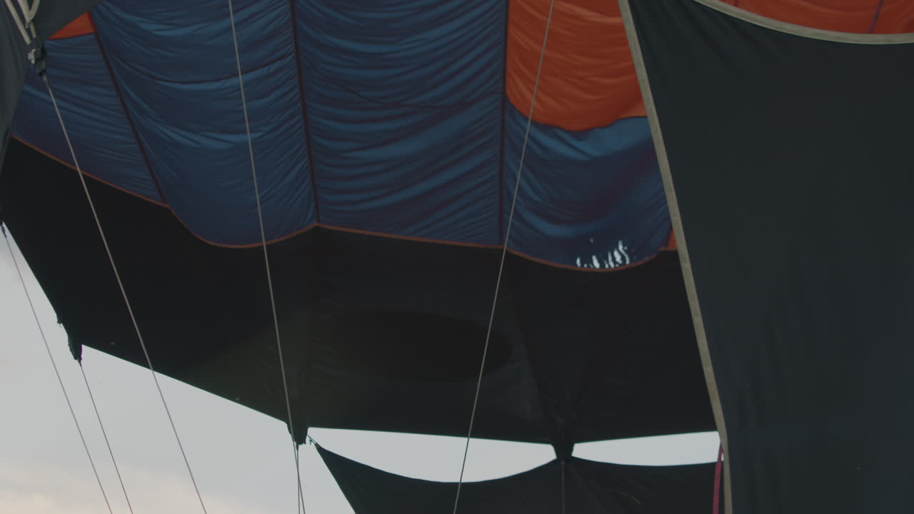 Underside view of vibrant hot air balloon envelope inflating as burner fire intensifies, sending orange flames upward through black opening, ropes tightening against overcast sky