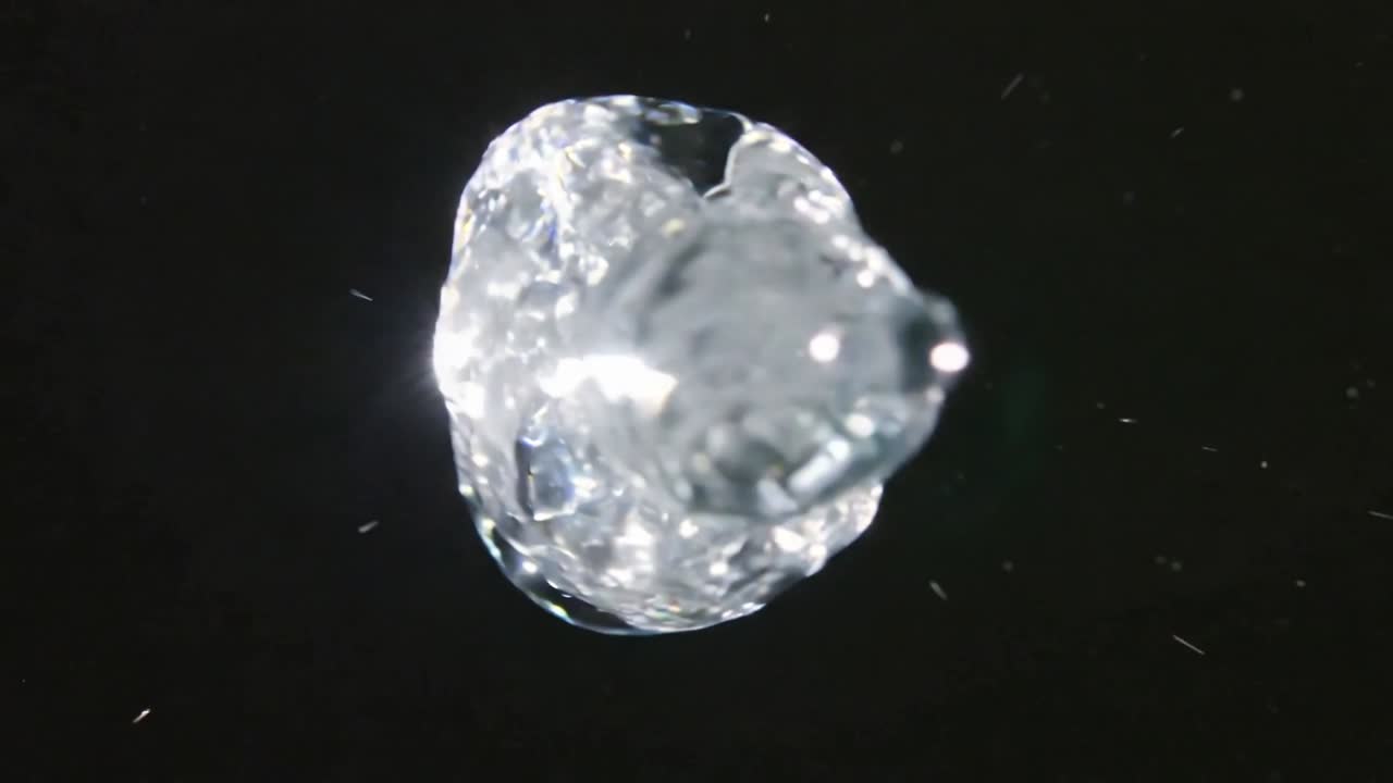 Una goccia simile al vetro che si deforma e si increspa sotto un getto di luce adeguato su uno sfondo nero, espellendo particelle.