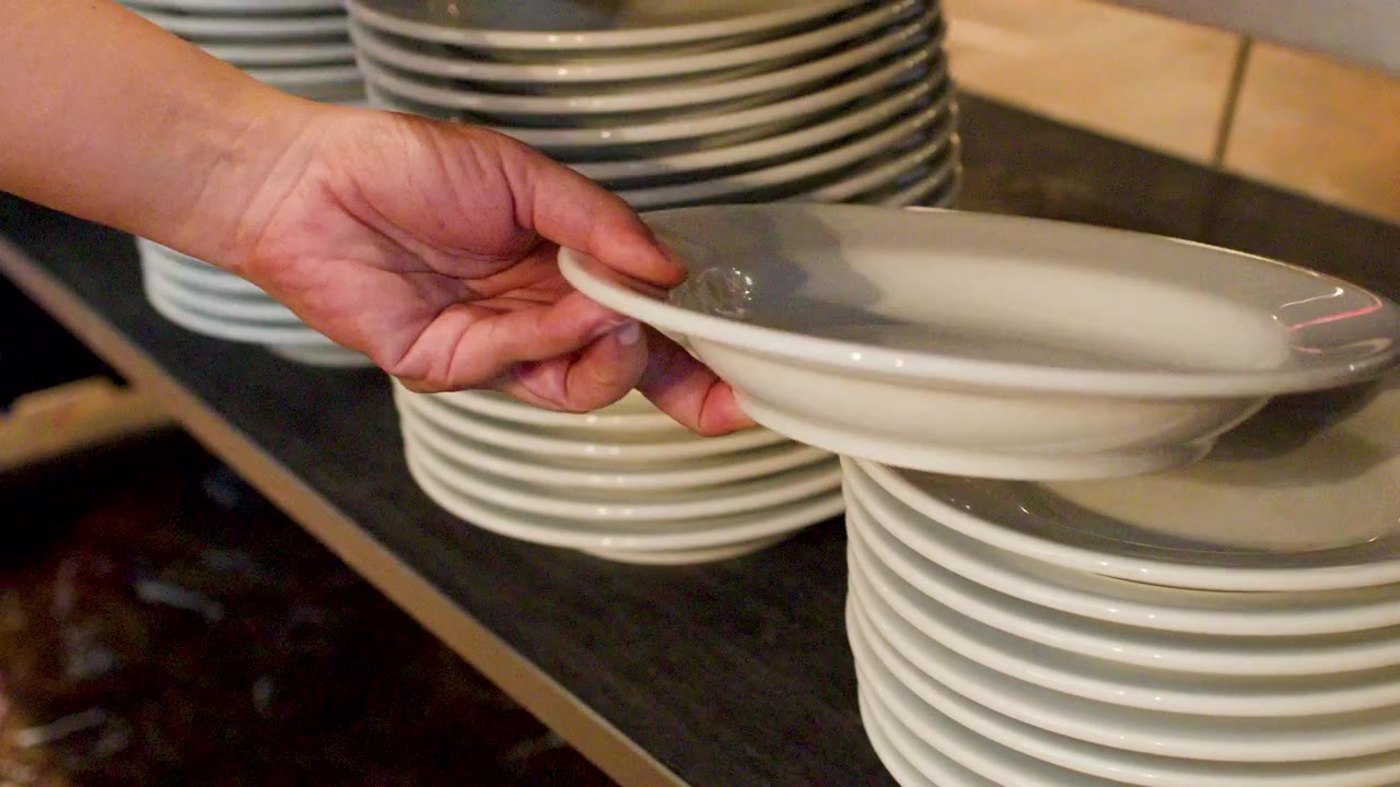 A person picks up clean white ceramic plates from a stack in a restaurant