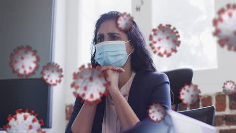 animation of covid 19 cells floating over worried biracial woman wearing face mask in office