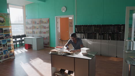 college student in casual clothing and cap sitting at desk in bright library, writing notes from open book surrounded by multiple stacked books, sunlight streaming through large window