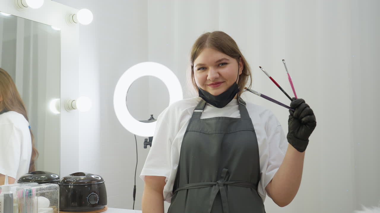 Beautician in dark apron and black gloves holds three makeup brushes while smiling at camera. Mirror reflection and salon tools are visible in bright, clean interior with soft lighting
