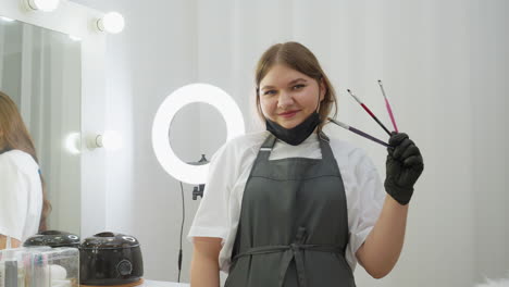 beautician in dark apron and black gloves holds three makeup brushes while smiling at camera. mirror reflection and salon tools are visible in bright, clean interior with soft lighting