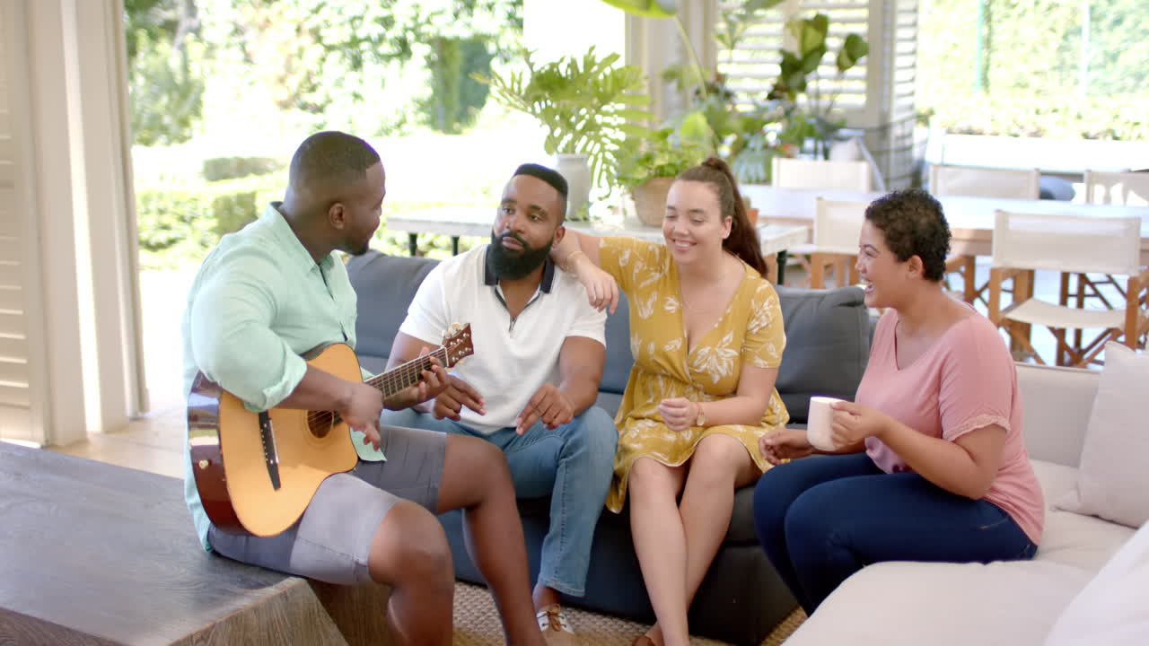 Playing guitar, african american man entertaining diverse friends sitting on couch together
