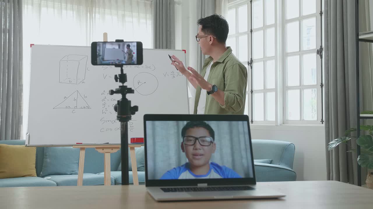 Asian Male Teacher With Glasses Shoots Video By Smartphone Camera And Having Video Call On Laptop While Teaching Math At Home