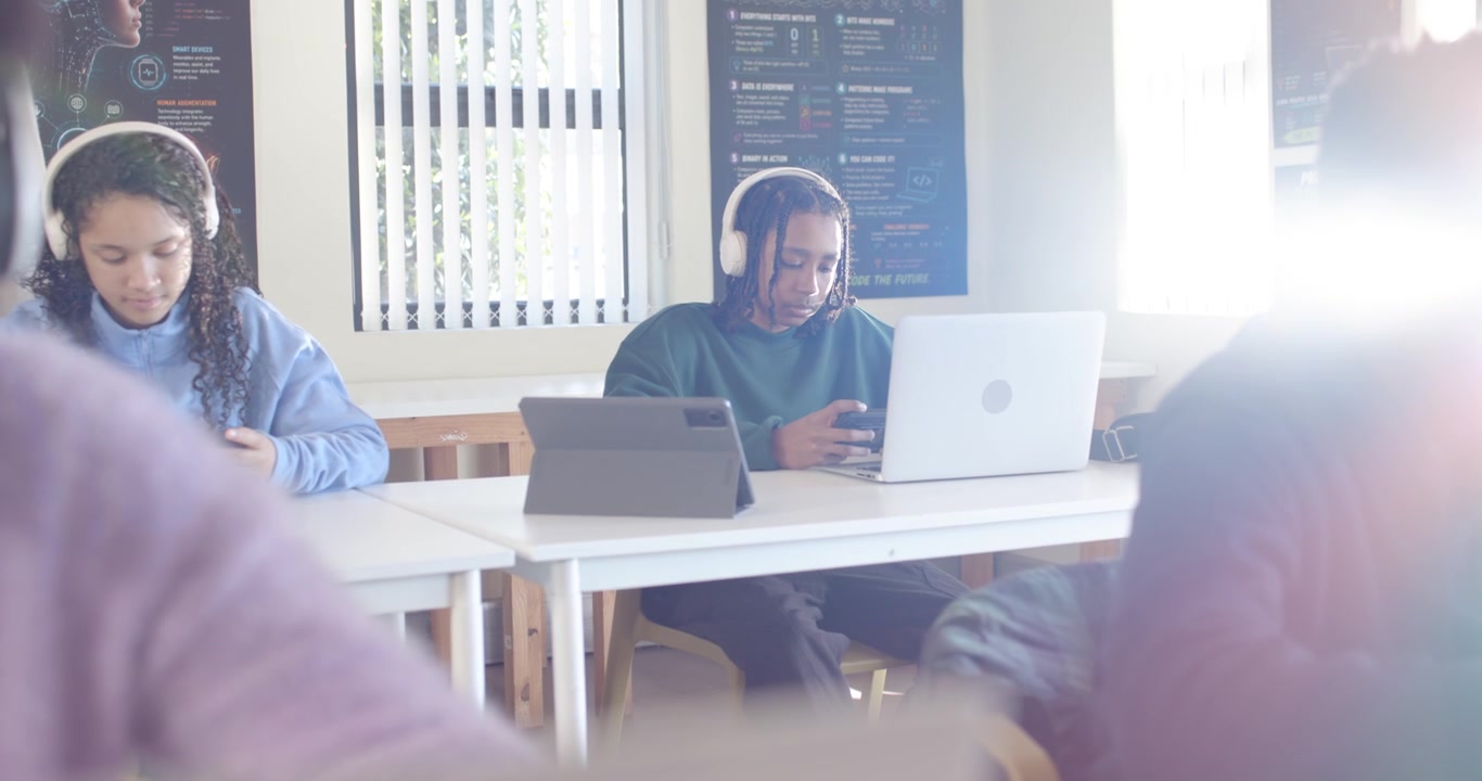 Diverse teens, male teen in green shirt scrolling smartphone, glancing at laptop for class at desks