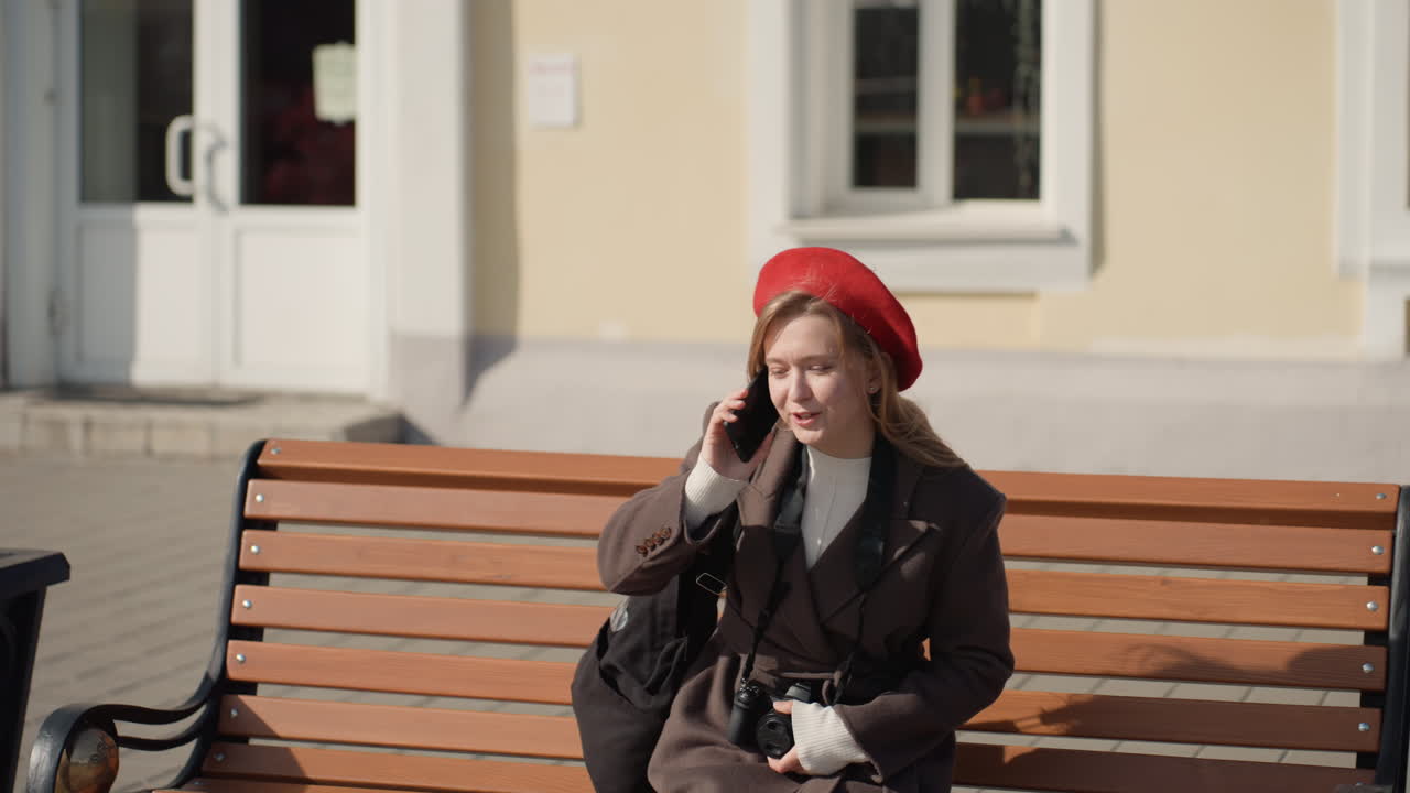 Kaukasisk kvinde nærmer sig en bænk, taler i telefon, iført rød baret og brun frakke, kamerataske ved siden, solrig gårdfacade, due i nærheden. Den rejsende holder pause, tjekker beskeder og sætter sig afslappet på bænken.