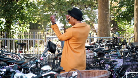 Joven-Negra-Con-Abrigo-Amarillo-Desabrochado,-Sombrero-Negro-Y-Gafas-De-Sol-Estacionando-Su-Bicicleta,-Poniéndose-Un-Bolso-Cruzado-Y-Alejándose,-Fuera-De-Tiro,-Sonriendo,-Vista-Lateral