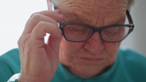 elderly woman adjusting glasses with her hand, focusing on reading, looking slightly frustrated while touching her glasses, seated indoors with bright natural light in the background