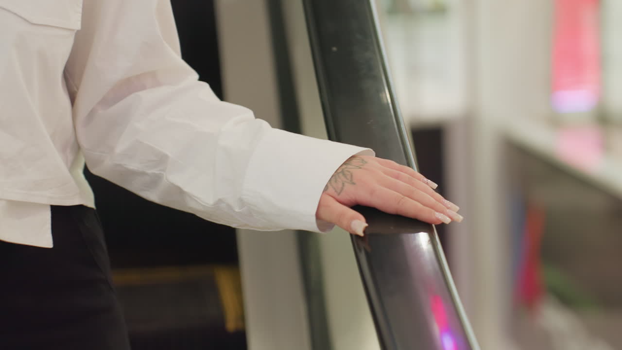 Close up of woman in white shirt and black pants holding escalator rail with tattooed hand while descending, illuminated by surrounding ambient lights in vibrant mall
