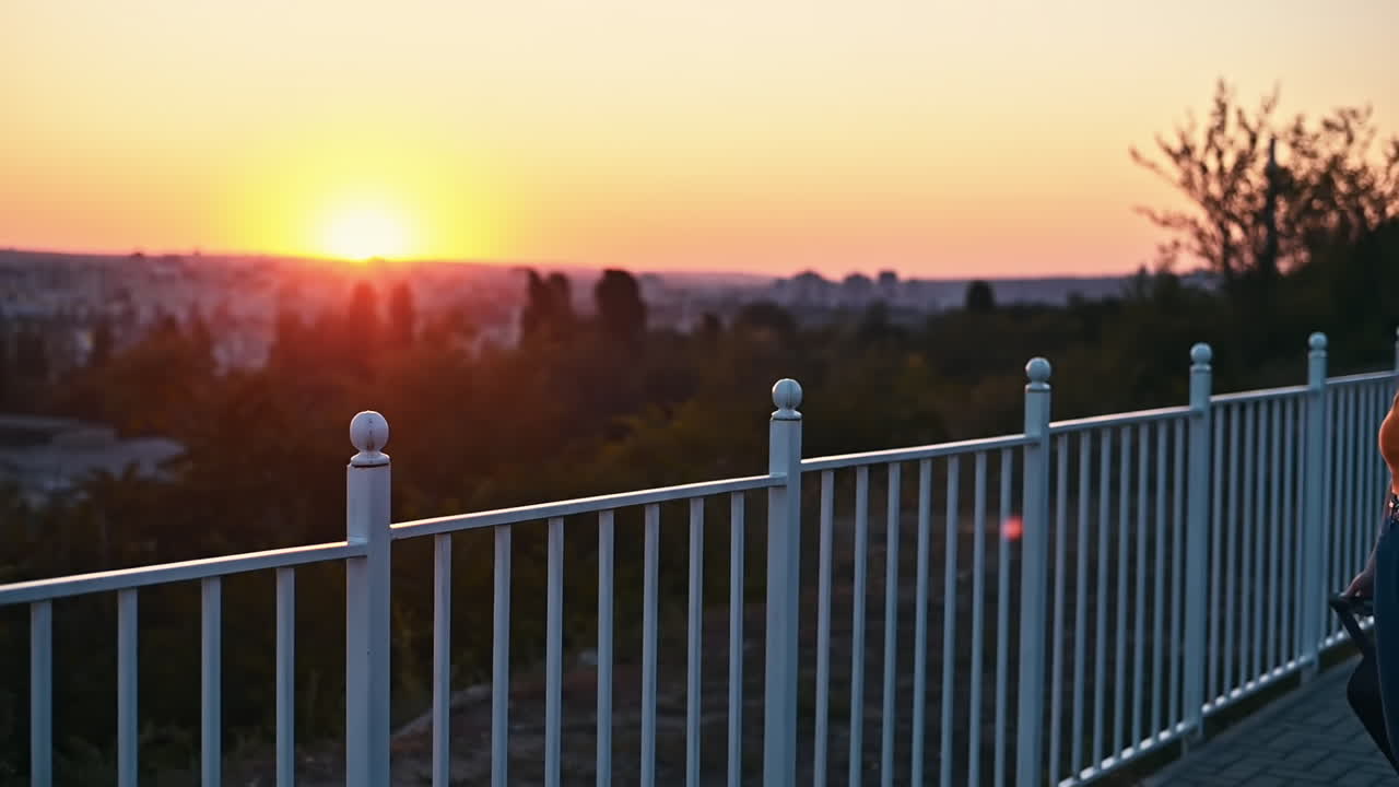 Young woman in carrot blouse looking at the city and enjoying orange sunset from the observation deck in the evening. Wearing protective medical mask. Rear view. Corona Virus idea