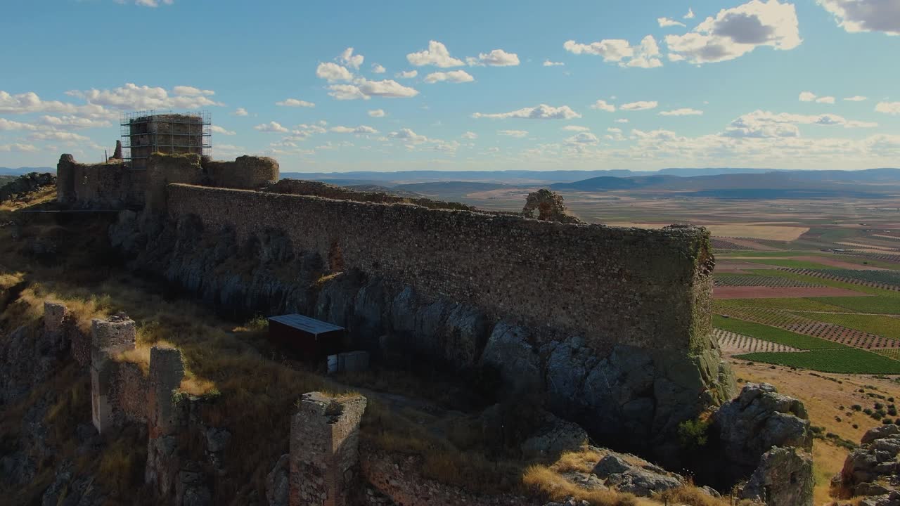 Un volo ascendente di un drone si inclina verso il basso rivelando la forma allungata del Castello di Peñas Negras, la sua posizione in cima alla collina e i circostanti uliveti, con lavori di restauro visibili sulla fortezza