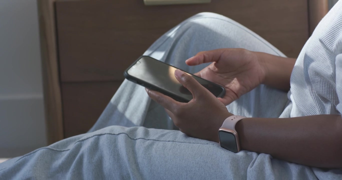 Dospělá Afroameričanka prochází telefon, spouští si ho na klín a usazuje se k odpočinku doma s hodinkami.