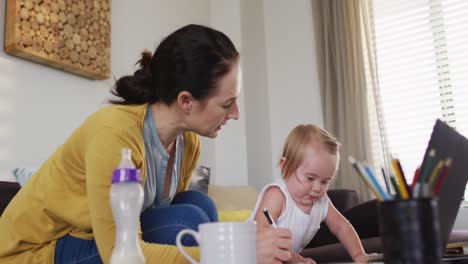 madre caucásica sosteniendo a su bebé tomando notas mientras trabaja desde casa