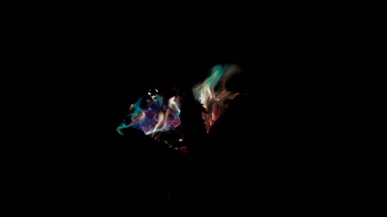 Abstract close-up footage of colorful flames and wisps of smoke burning in darkness on a black background, glowing teal, purple, and orange for dramatic texture