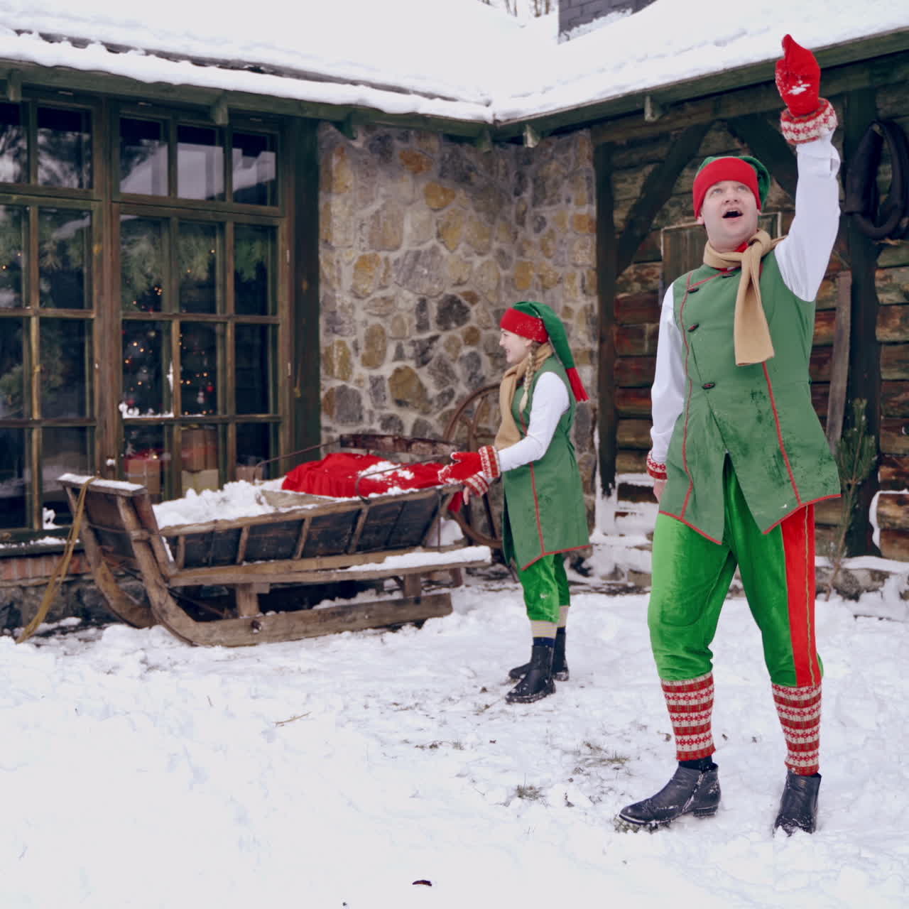 Two happy elves playing snowballs outdoors. Santa Claus looking through the window from the house. Christmas time.