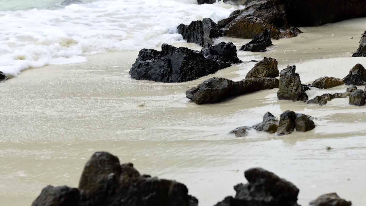 Close-up view of waves softly lapping against dark rocks on a sandy beach.