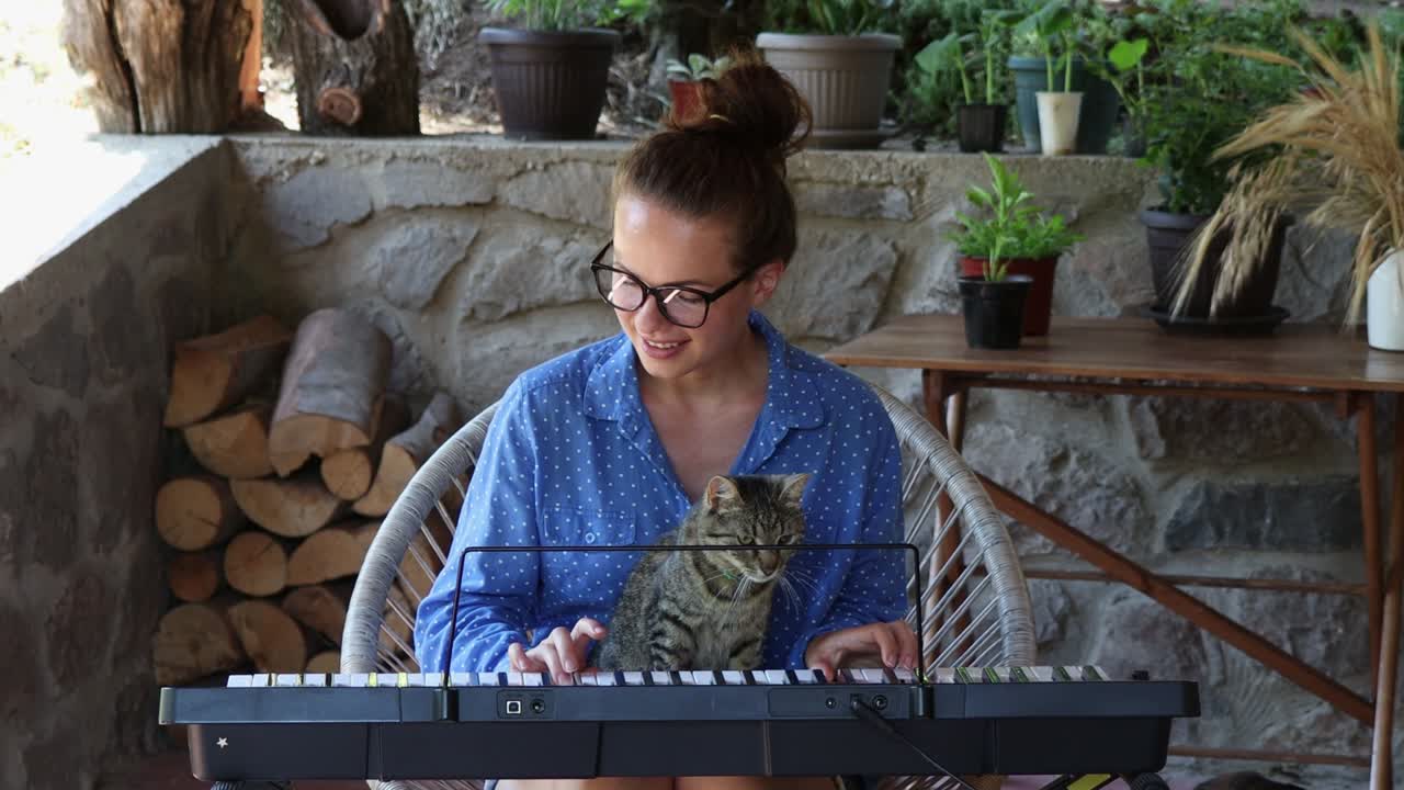 joven feliz con gafas sentada junto con su lindo gato y tocando el piano eléctrico.