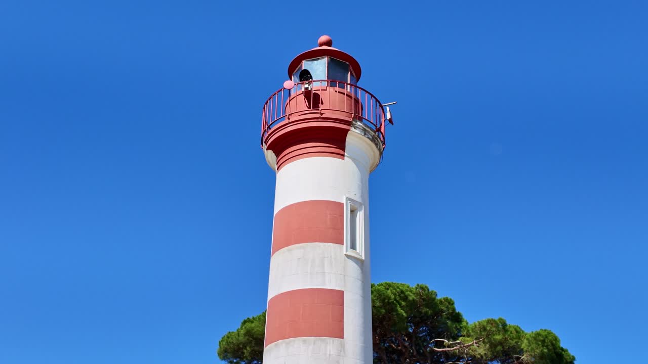 Pullback aerial drone shot of La Rochelle Old Port lighthouse with its red and white stripes, framed by a clear blue sky and green treetops - France