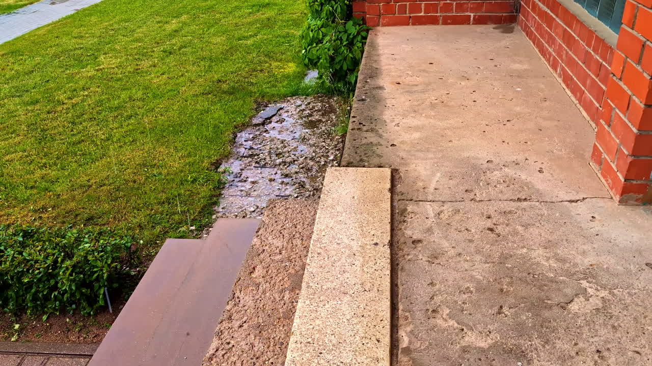 Concrete porch with rain falling and droplets in puddle near brick house and lawn