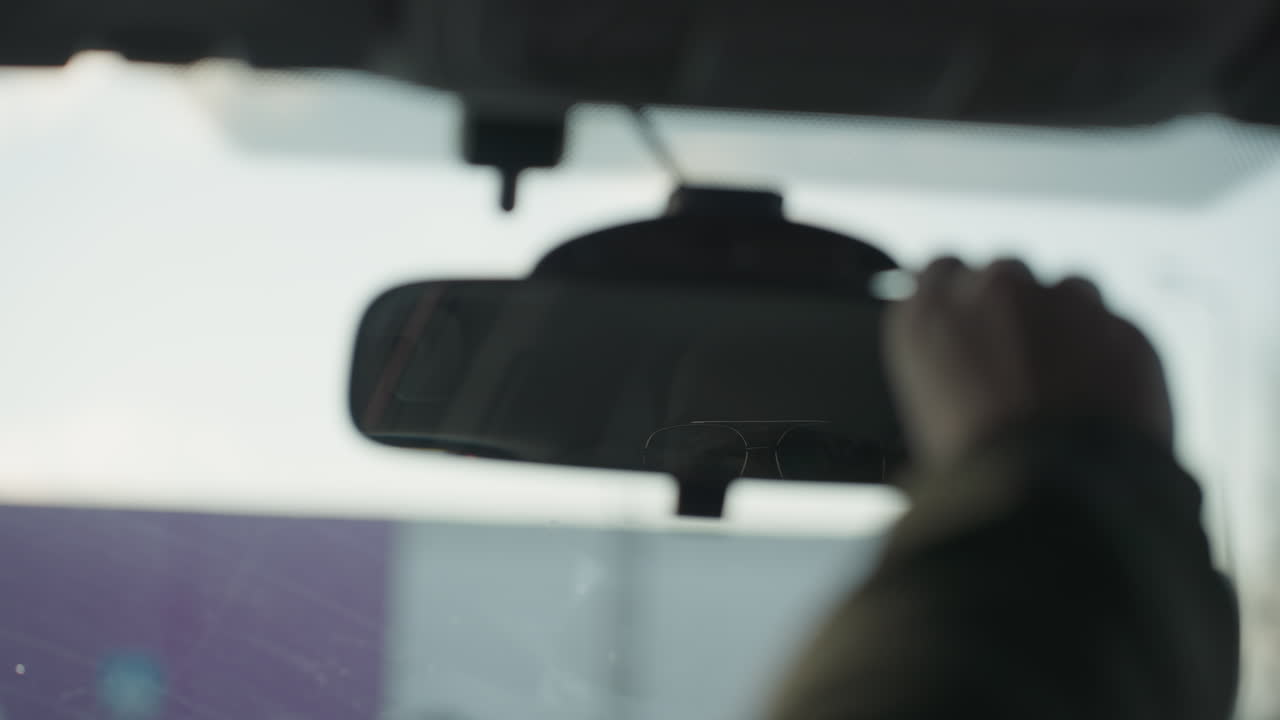 rear view mirror reflecting young driver wearing glasses adjusting mirror upward with hand inside parked car with soft focus winter background and diffused daylight through windshield