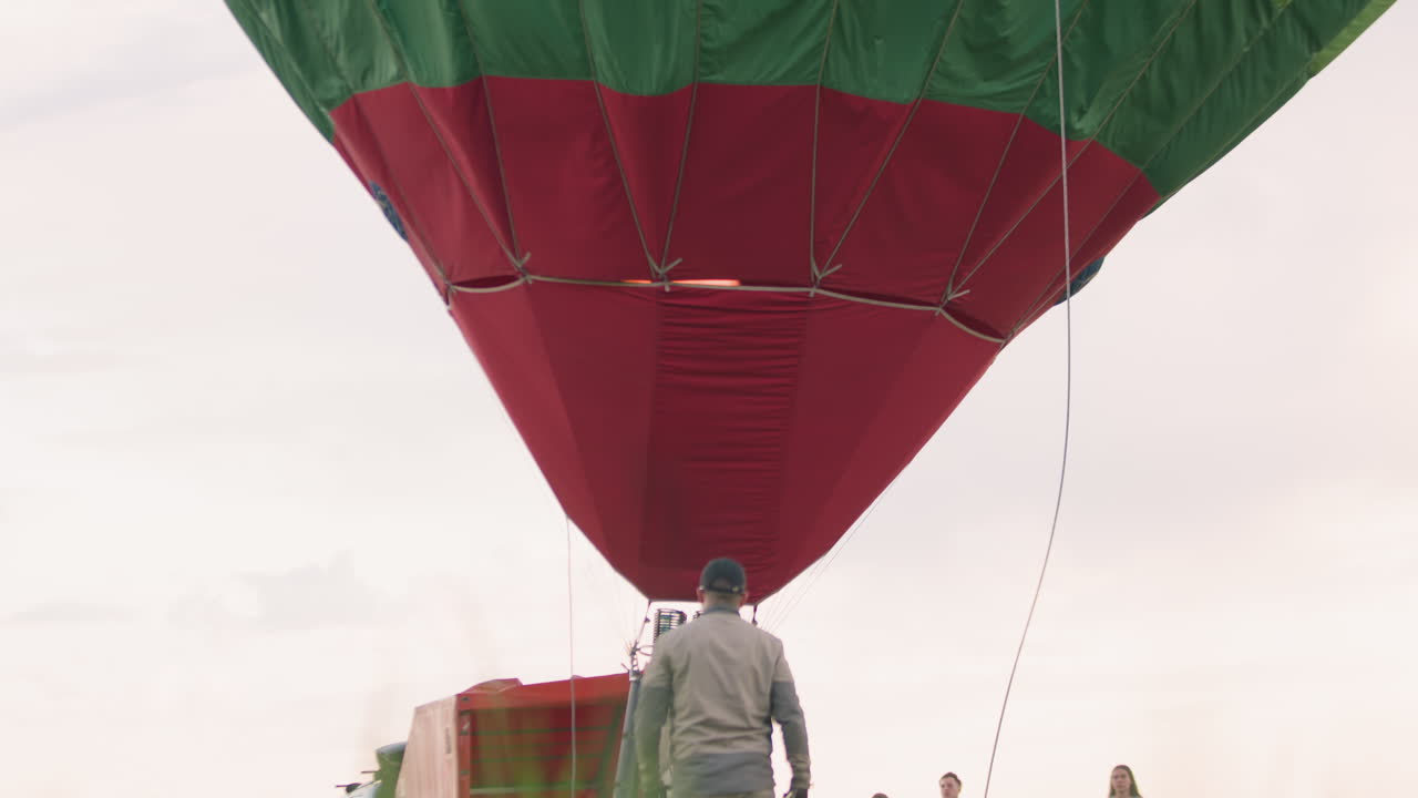 Back view of man walking toward colorful hot air balloon envelope while crew stand nearby on green field under pastel evening sky, vibrant panels stretching overhead