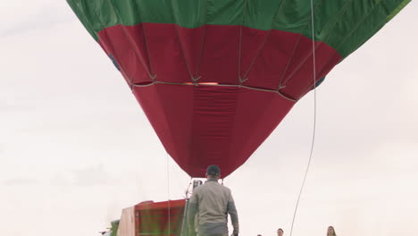 back view of man walking toward colorful hot air balloon envelope while crew stand nearby on green field under pastel evening sky, vibrant panels stretching overhead