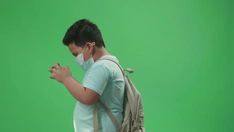 side view of asian boy student wearing a mask and playing video game on mobile phone while walking to school on green screen chroma key