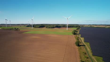 panning aerial view passing across sustainable solar panel array and wind turbines on rural farmland