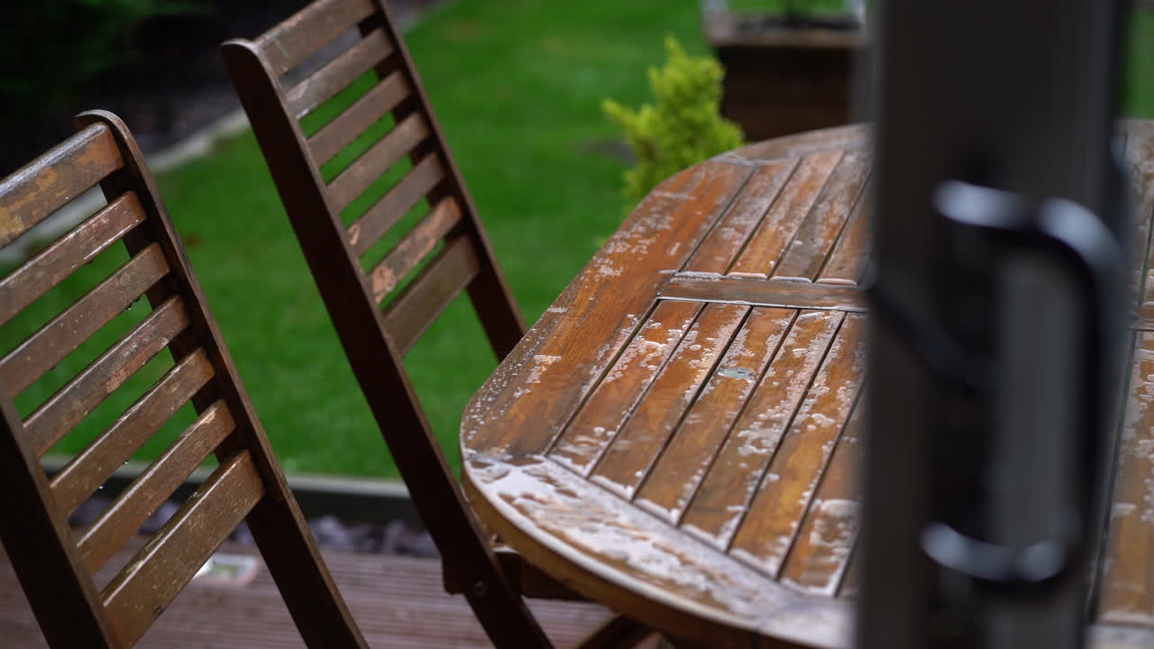 Regn på et træhavebord gennem en dør med træstole og vådt grønt græs