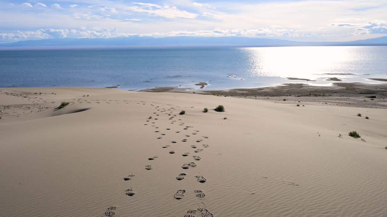 A trail of footprints marks a solitary journey down a vast sand dune to the sparkling shores of Durgun Nuur lake. A scene of reaching a remote and beautiful destination