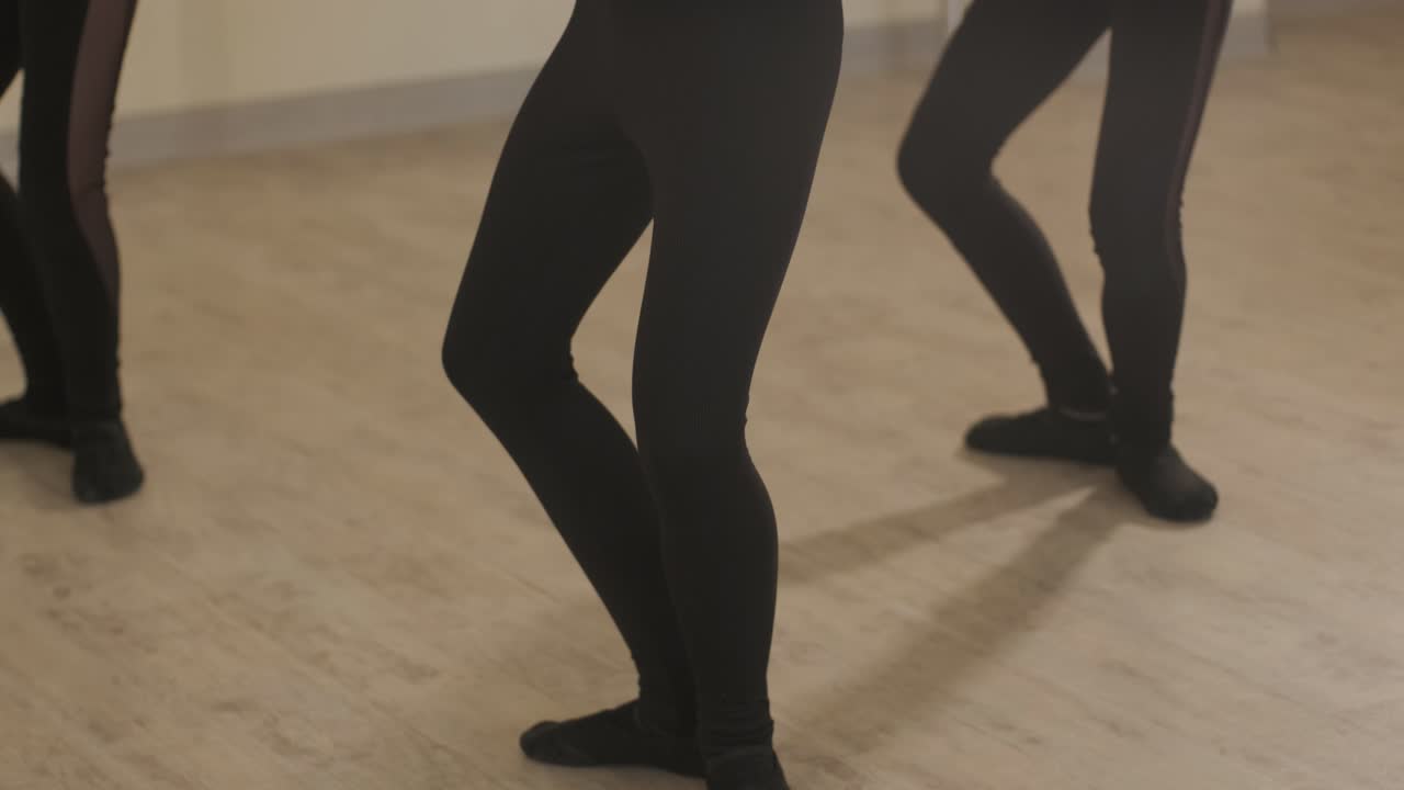 A group of young ballet students in black dancewear practicing positions in a spacious ballet studio with wooden flooring and wall-mounted barres. Focused expressions and synchronized movements.