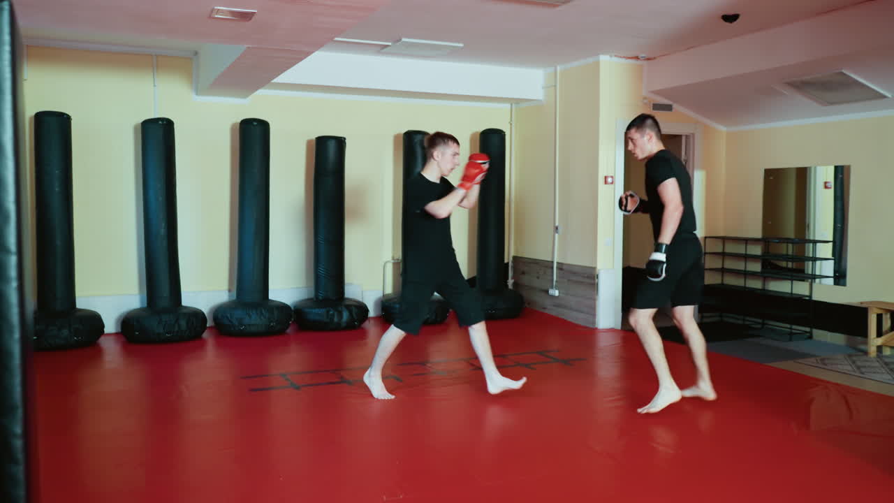 Wrestlers sparring inside gym, wearing gloves and black outfits, preparing for combat sport practice with focused stance and movement on red mat floor surrounded by standing punching bags during martial training