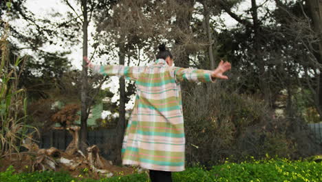 woman in plaid coat dances near a garden with trees and yellow flowers in spring