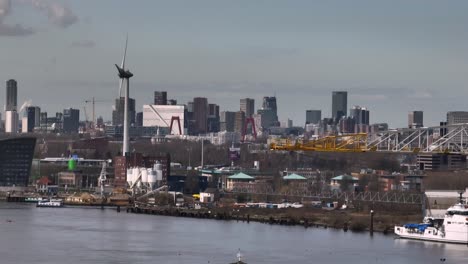 Aerial-from-behind-cranes-over-Nieuwe-Maas-river-of-bank-with-turbine,-Rotterdam