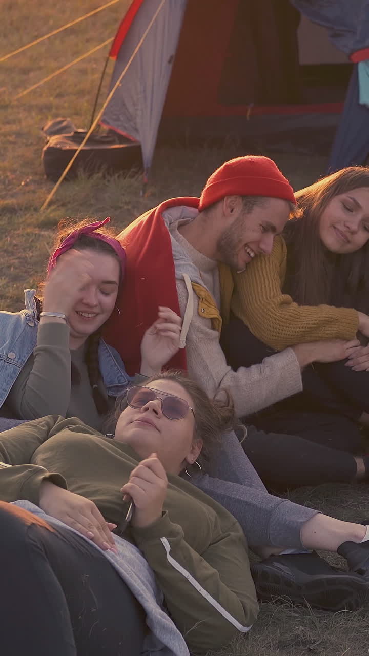 group of positive young tourists rests at burning bonfire in camp with blue tent in warm autumn evening at sunset