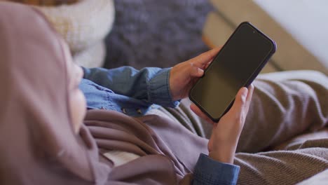 High-angle-video-of-biracial-woman-in-hijab-sitting-at-home-using-smartphone,-copy-space-on-screen