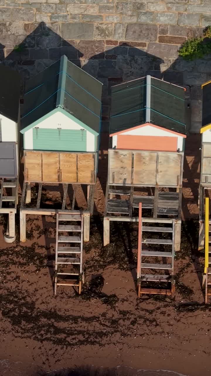 Vertical aerial drone pans across Torquay beach huts. View highlights colorful doors and rooftops above sandy beach. Perfect seaside vacation stock clip with holiday, leisure, and relaxation themes