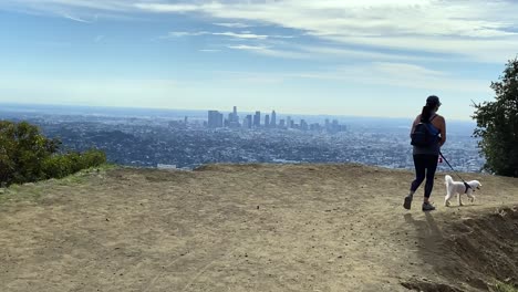 Female-hiking-with-dog-scenic-viewpoint-overlooking-hazy-downtown-Los-Angeles-city-skyline