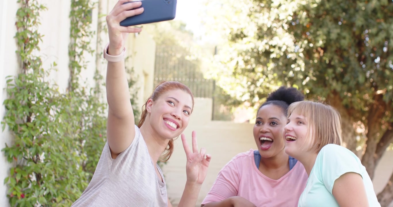 Tre donne in abbigliamento sportivo sui tappetini da yoga, la donna a sinistra che maneggia uno smartphone blu e posa per un selfie.
