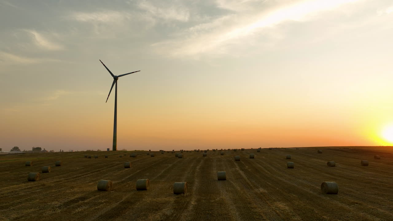 Wind turbine and round hay bales on farmland at golden sunset, renewable rural power