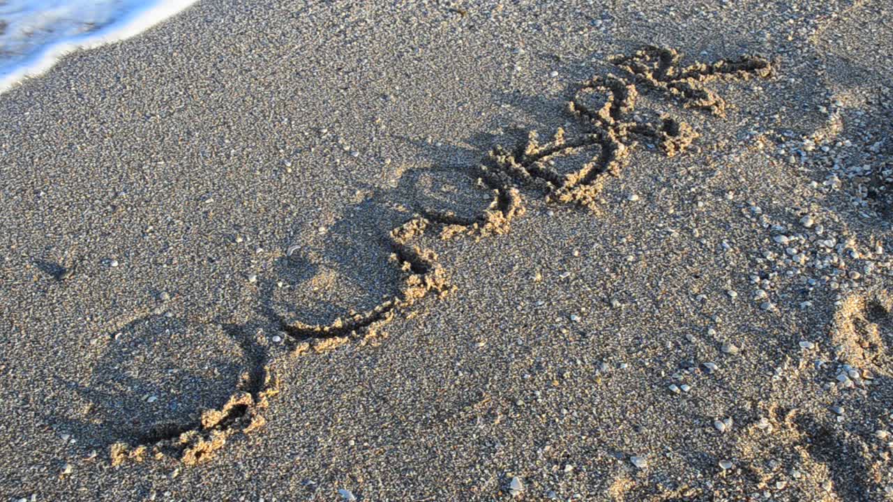 Tulisan 'Oktober' di pasir. Tulisan itu terhapus oleh ombak laut. Rekaman di pantai.